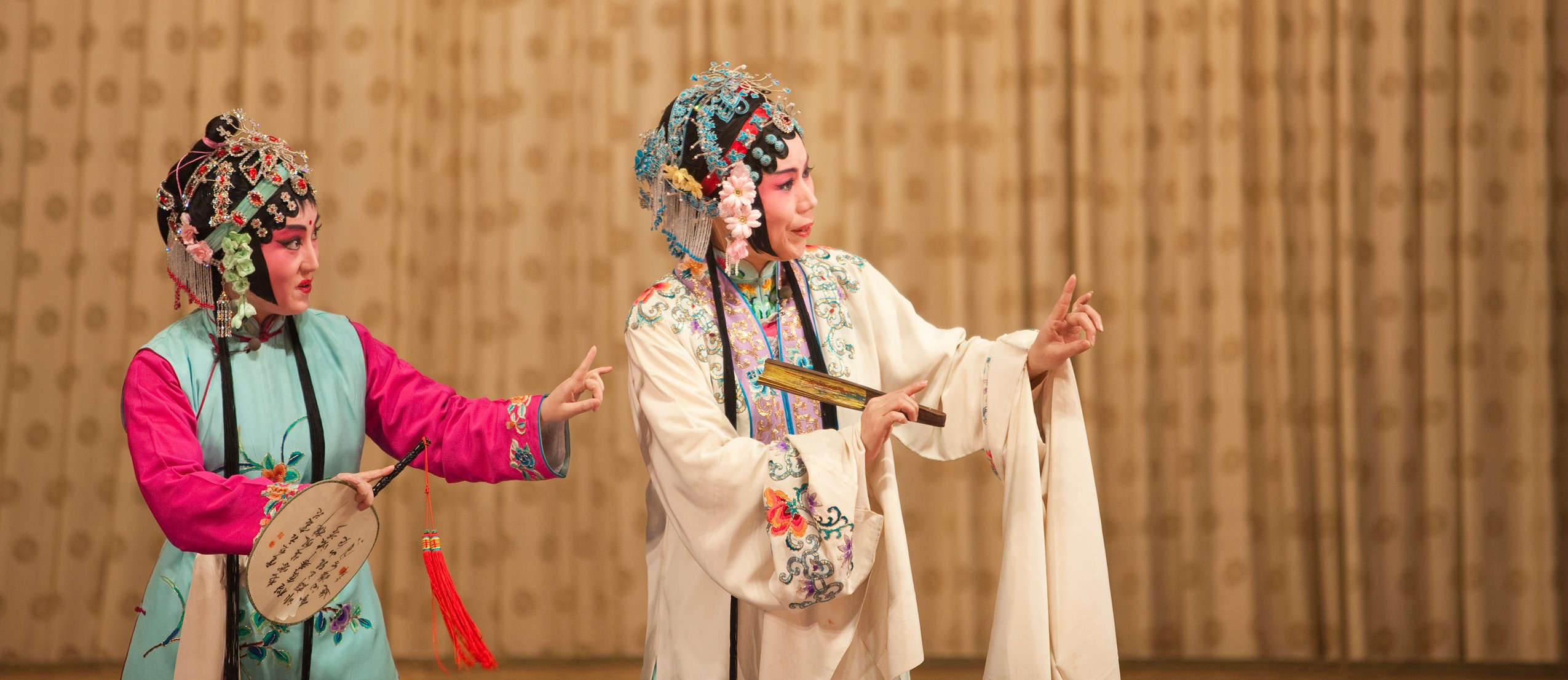 Two performers in elaborate Chinese opera costumes stand on stage against a patterned gold backdrop. 