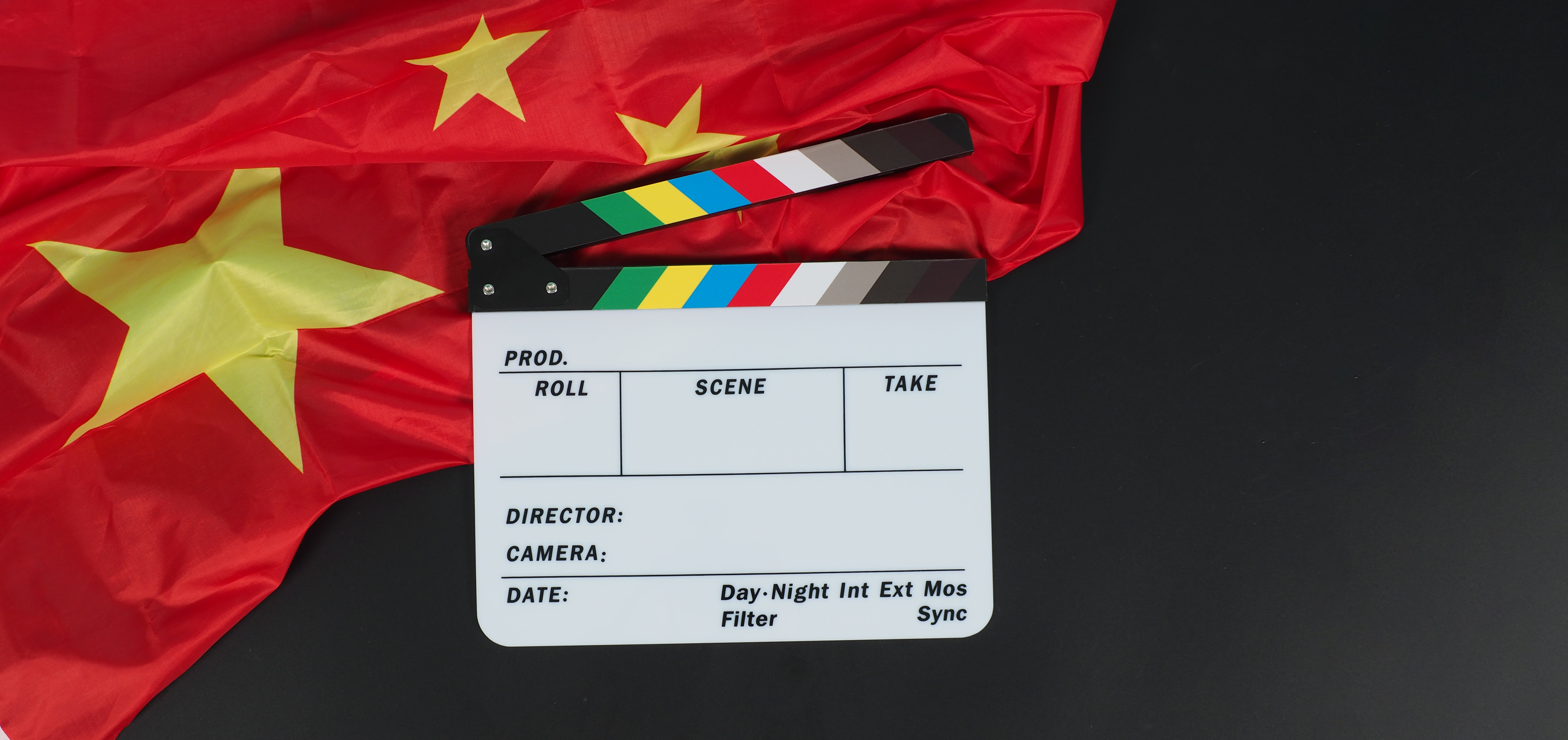 The image shows a movie clapperboard placed on a Flag of China. The clapperboard has labeled sections for production details, including fields for "Roll," "Scene," "Take," "Director," "Camera," and "Date," along with some colored stripes at the top.