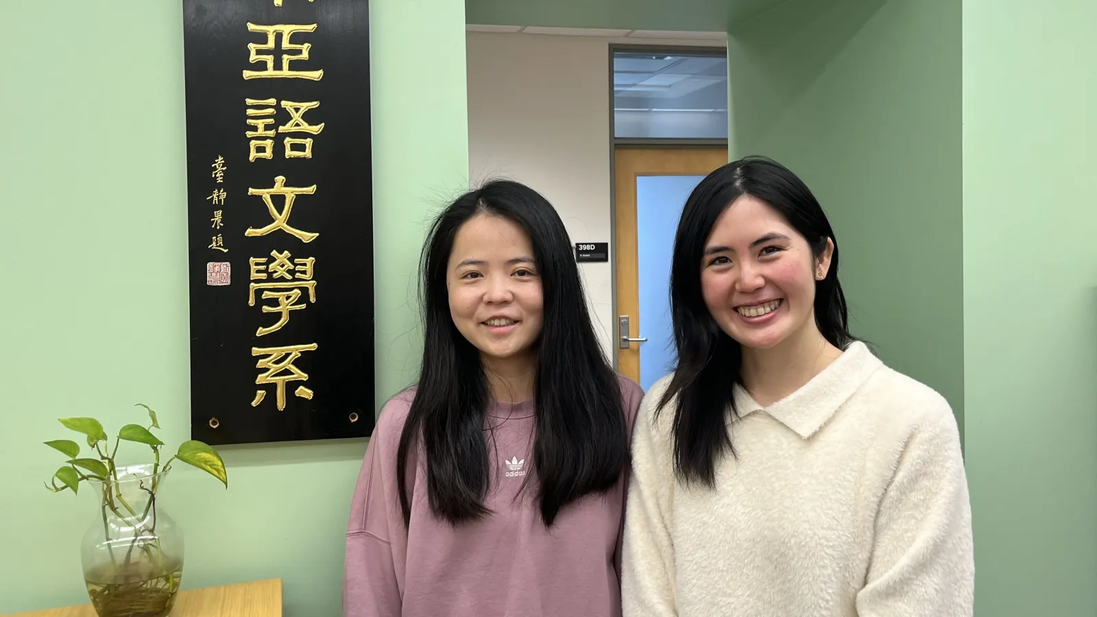 Two women standing in front of a green wall with a plant next to them