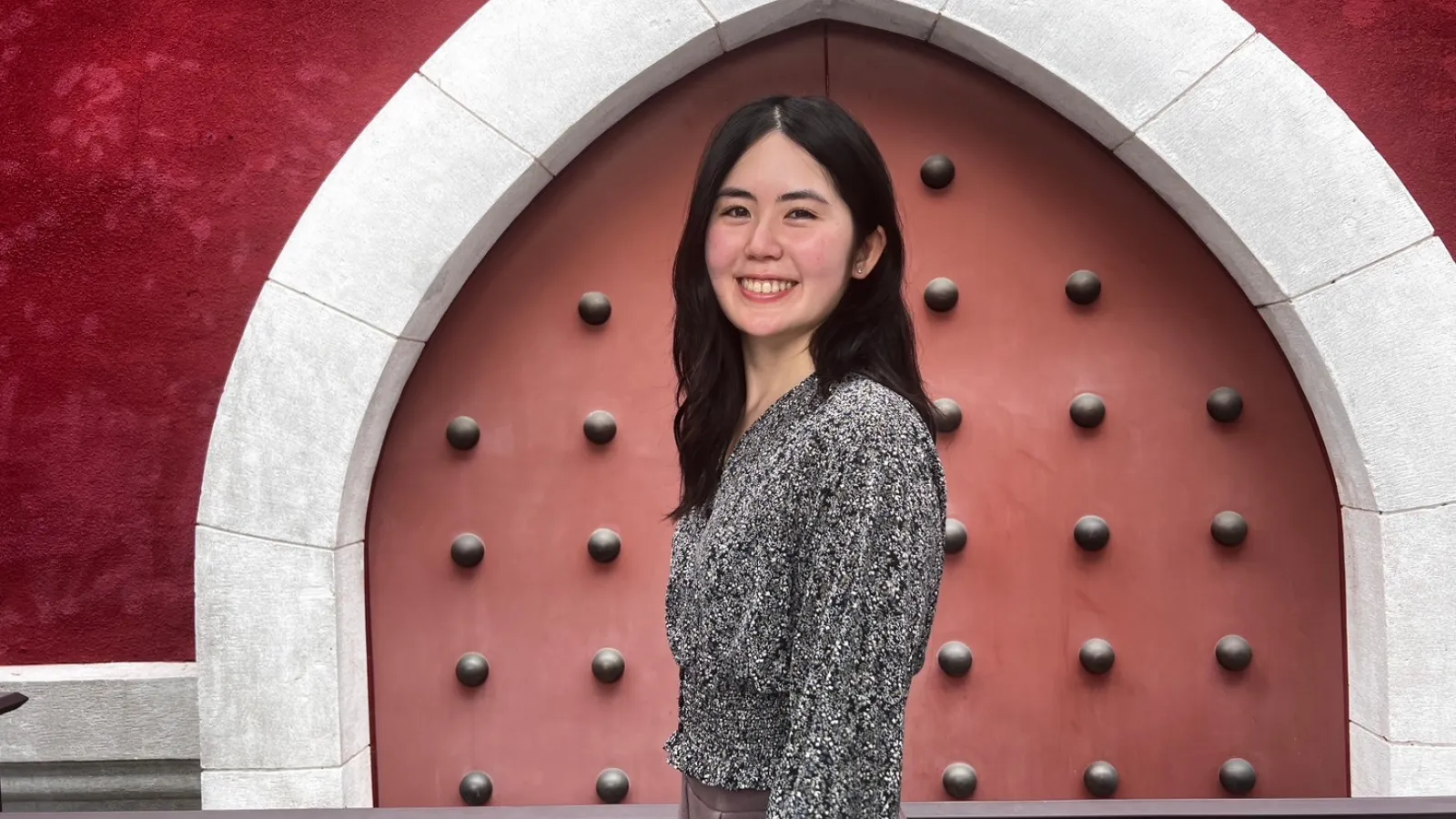 A woman smiles while standing in front of a large, red, studded door set in an archway with a dark red wall.