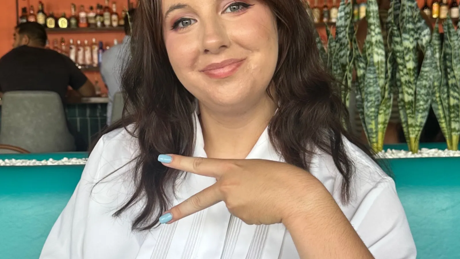  A woman with long brown hair and a white shirt sits at a restaurant table, smiling and making a peace sign with her hand. There are plants and bottles on shelves in the background. 
