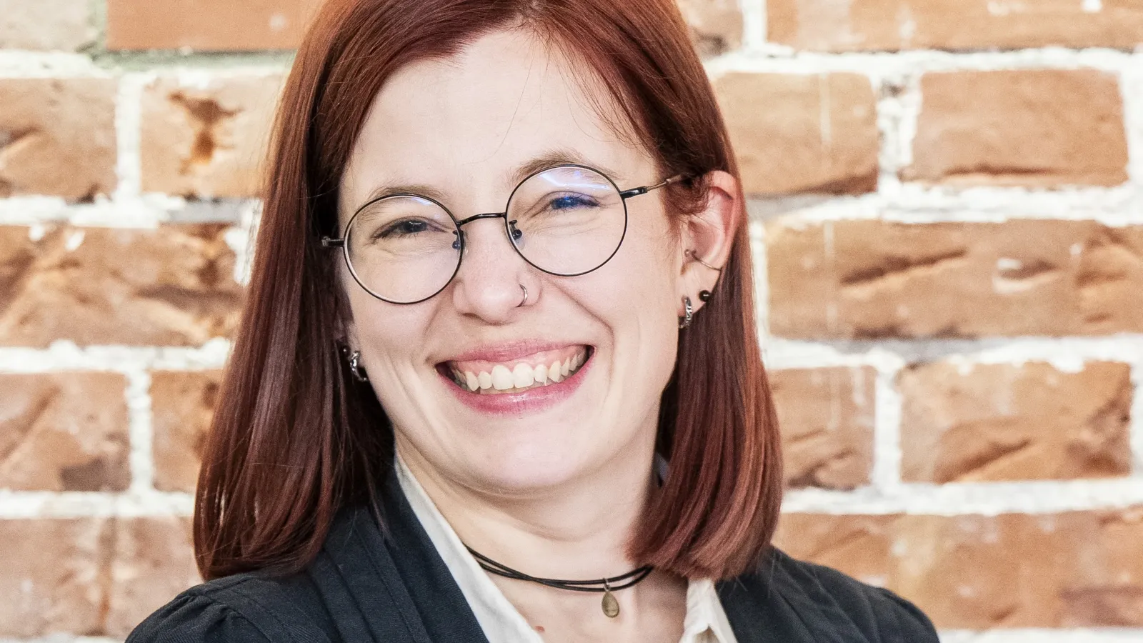 A woman with red hair and glasses smiles brightly against a rustic brick wall backdrop