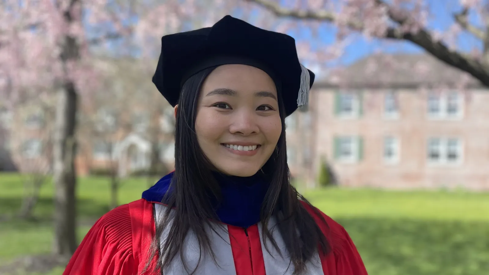 A woman dressed in a graduation gown and cap stands against a backdrop of a tall tree.  