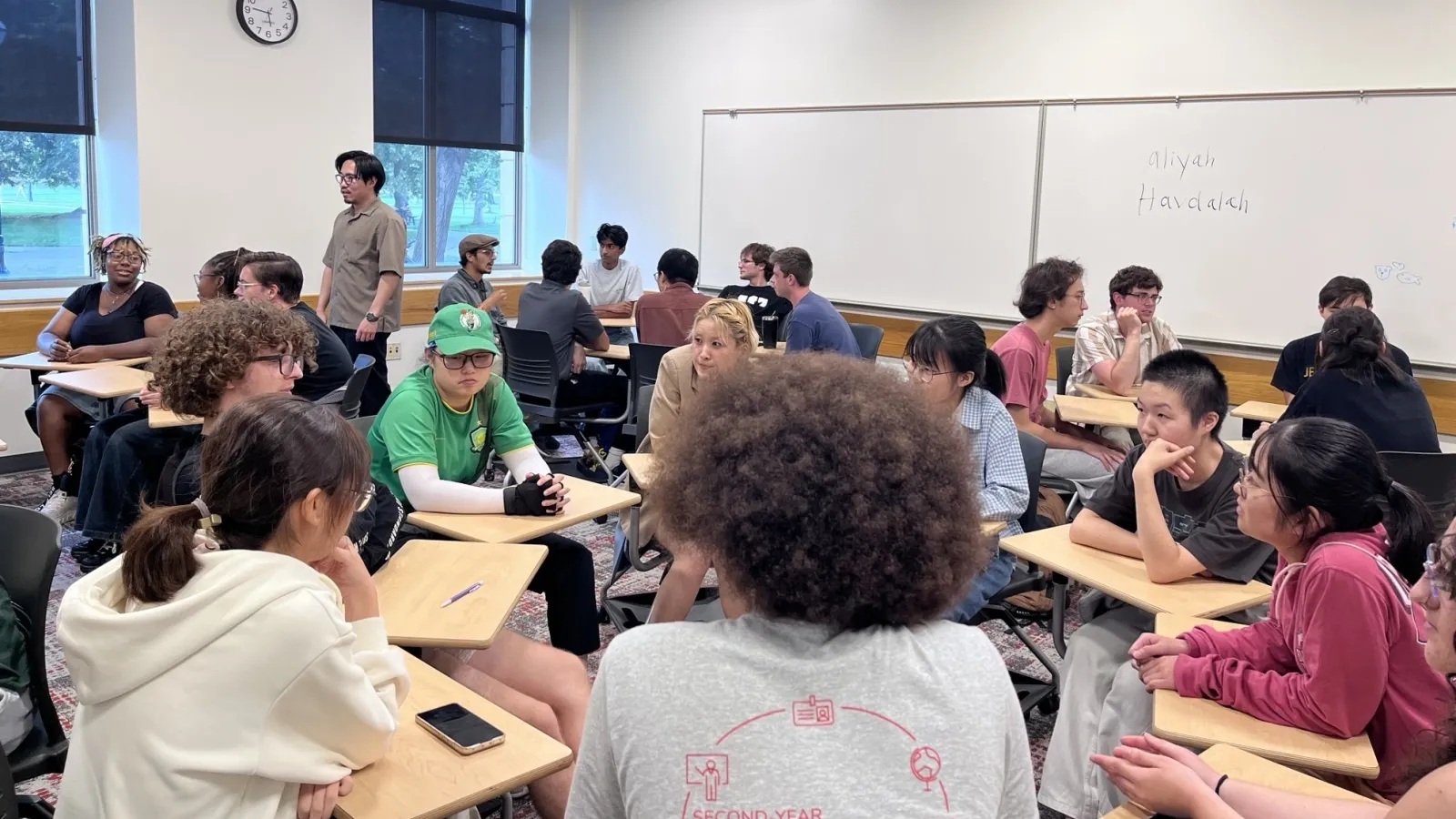 Students sit in small groups at desks in a classroom, engaged in discussion. A person stands near the whiteboard, which has words written on it.