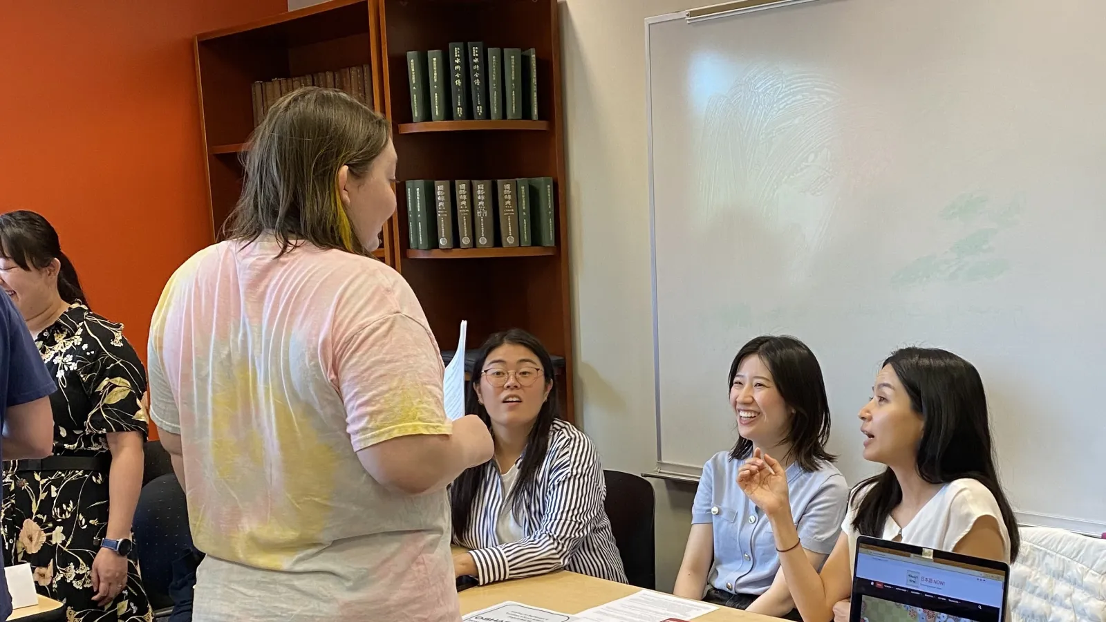  A person stands and talks to three seated individuals behind a table at an indoor event. The table displays name tags, papers, and a laptop. Other people are present in the background. 