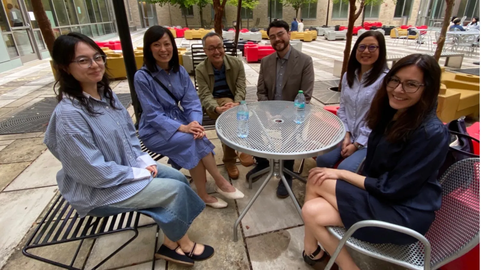  Six people sit around a circular metal table outdoors, smiling at the camera. The table has three water bottles, and the setting includes trees and modern buildings. 