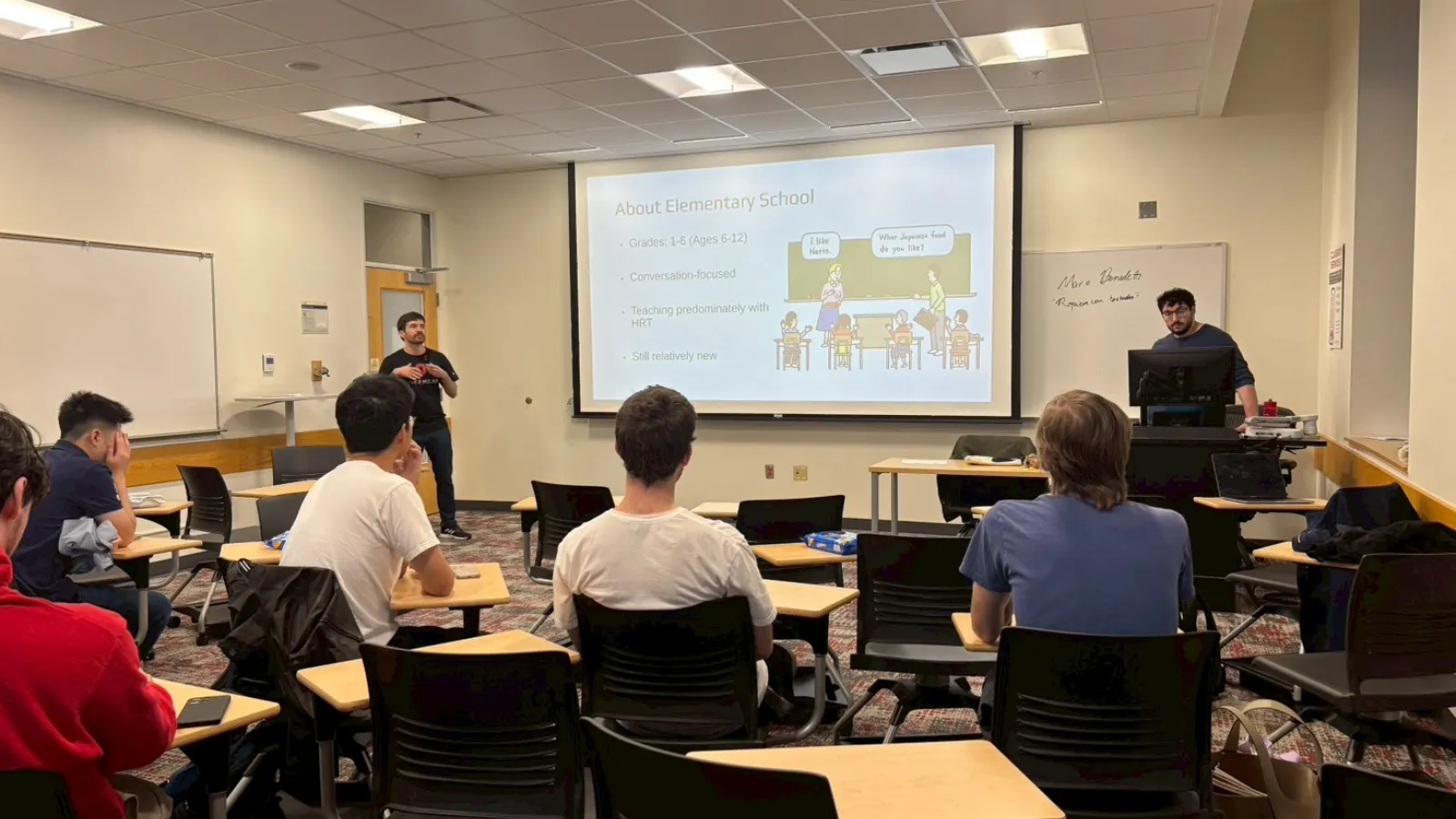 A classroom with students seated at desks facing two presenters; a slide about elementary school is projected on a screen at the front of the room. 