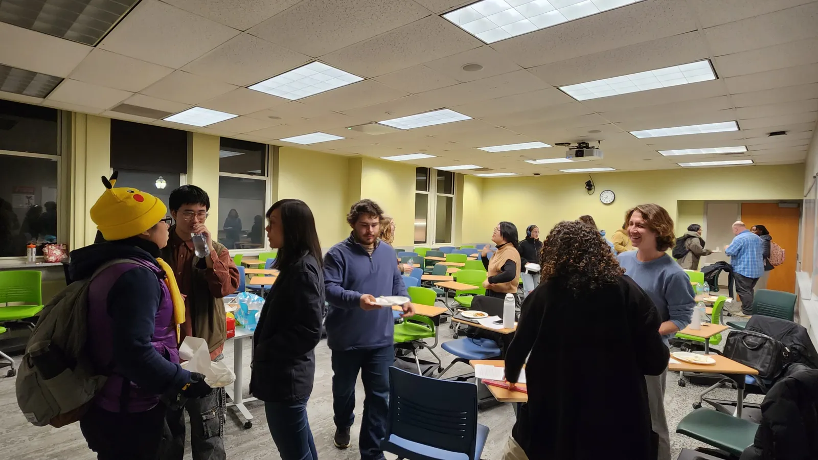  A group of people stand and talk in a classroom with green chairs and overhead lights. Some hold plates of food. The room appears to be set up for a casual gathering or event. 
