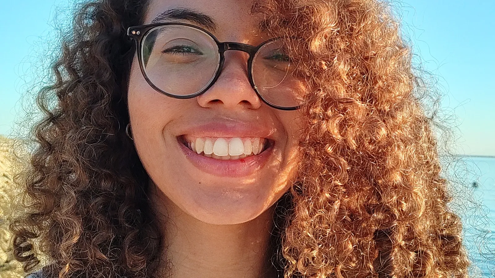 Justine Franklin, woman with long curly hair, a big smile, round frame glasses, in a t-shirt in the sunlight