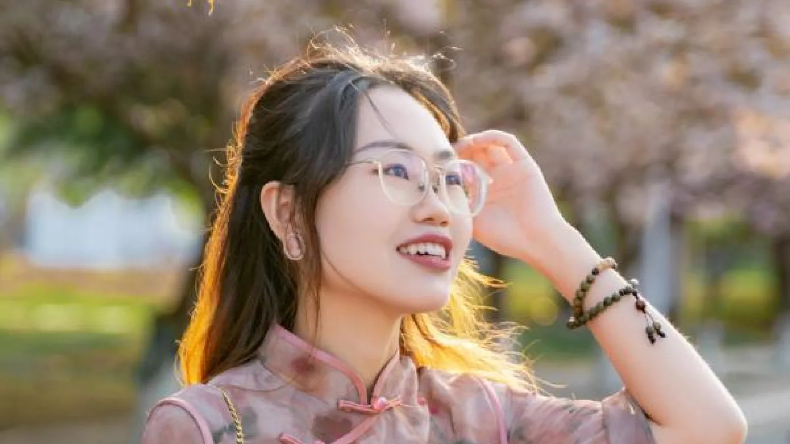 A woman wearing glasses and a floral dress stands in front of a tree, smiling and enjoying the outdoors.  