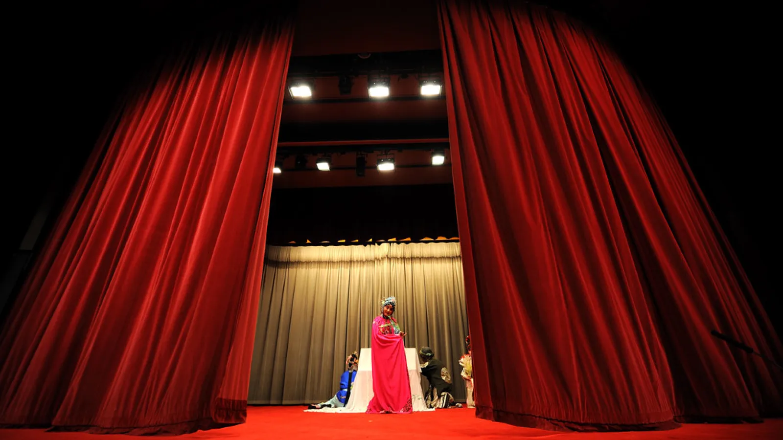 A theater stage with red velvet curtains drawn open, revealing a figure in colorful attire under stage lighting.