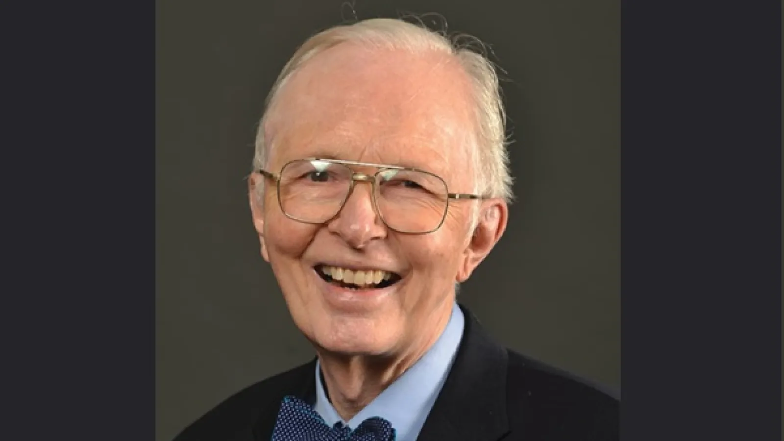  An elderly man with gray hair, glasses, and a blue bow tie smiles at the camera against a plain dark background. 