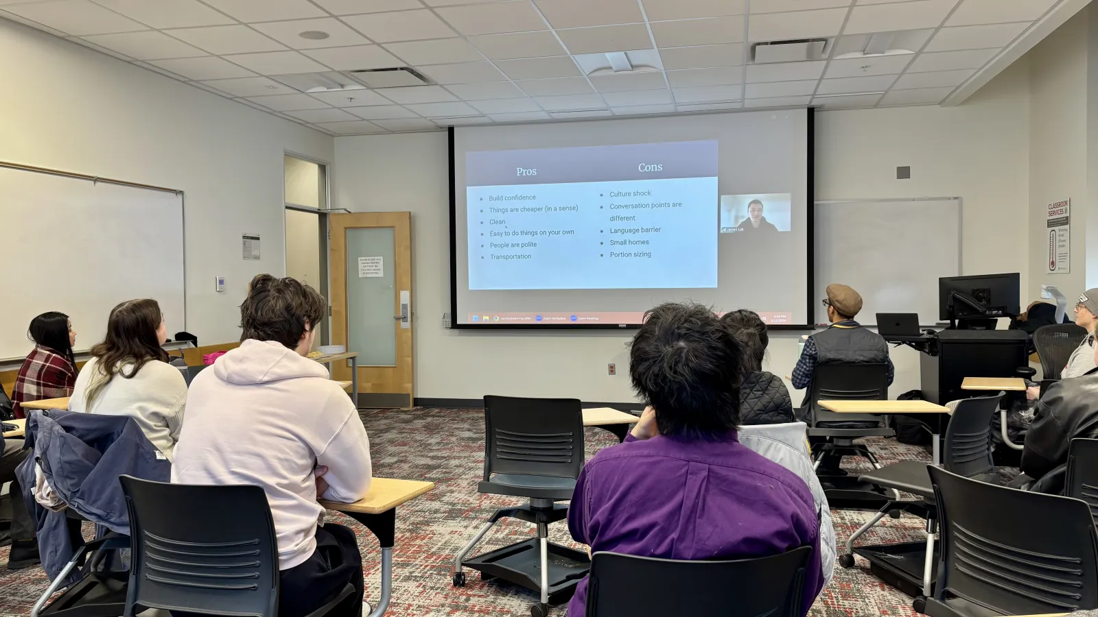 Students in a classroom facing a large screen showing a video call presentation. The room has desks, chairs, and a carpeted floor, creating a focused learning atmosphere.