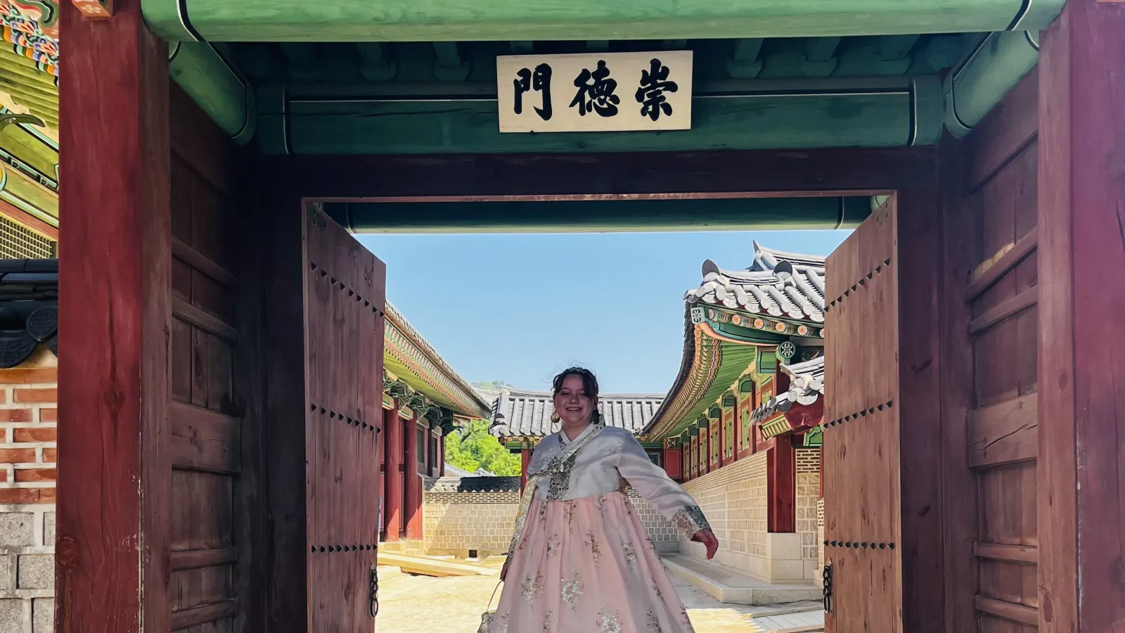  A young woman wearing traditional Korean hanbok stands under an open wooden gate at a historical Korean palace or temple on a sunny day. 