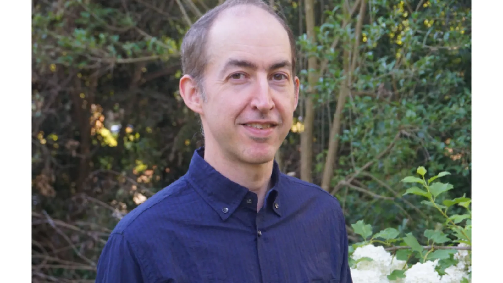 Picture of a man wearing a blue polo shirt with green foliage behind him