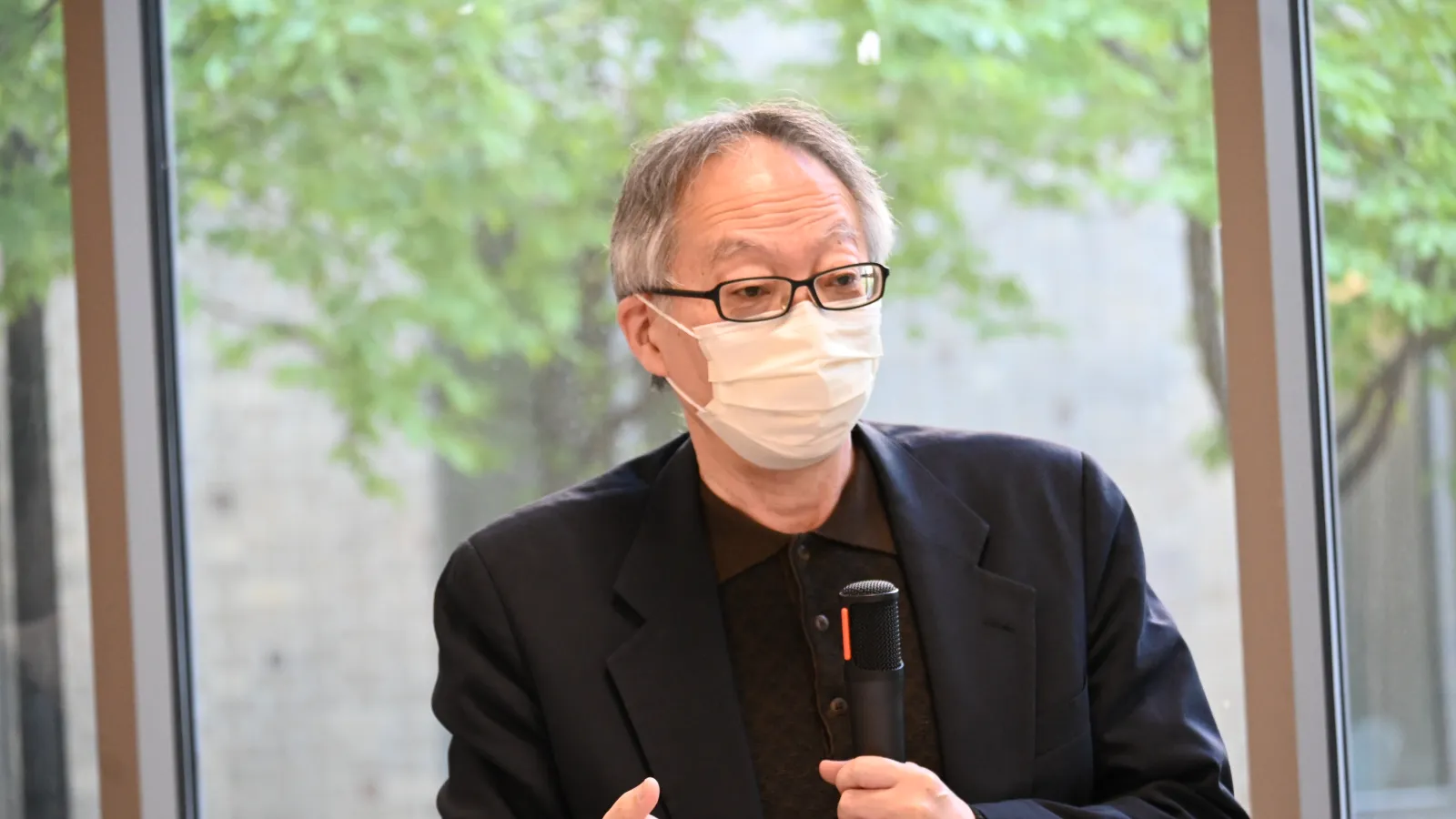  An older man wearing a face mask, glasses, and a suit jacket holds a microphone and gestures with one hand while speaking indoors near large windows. 