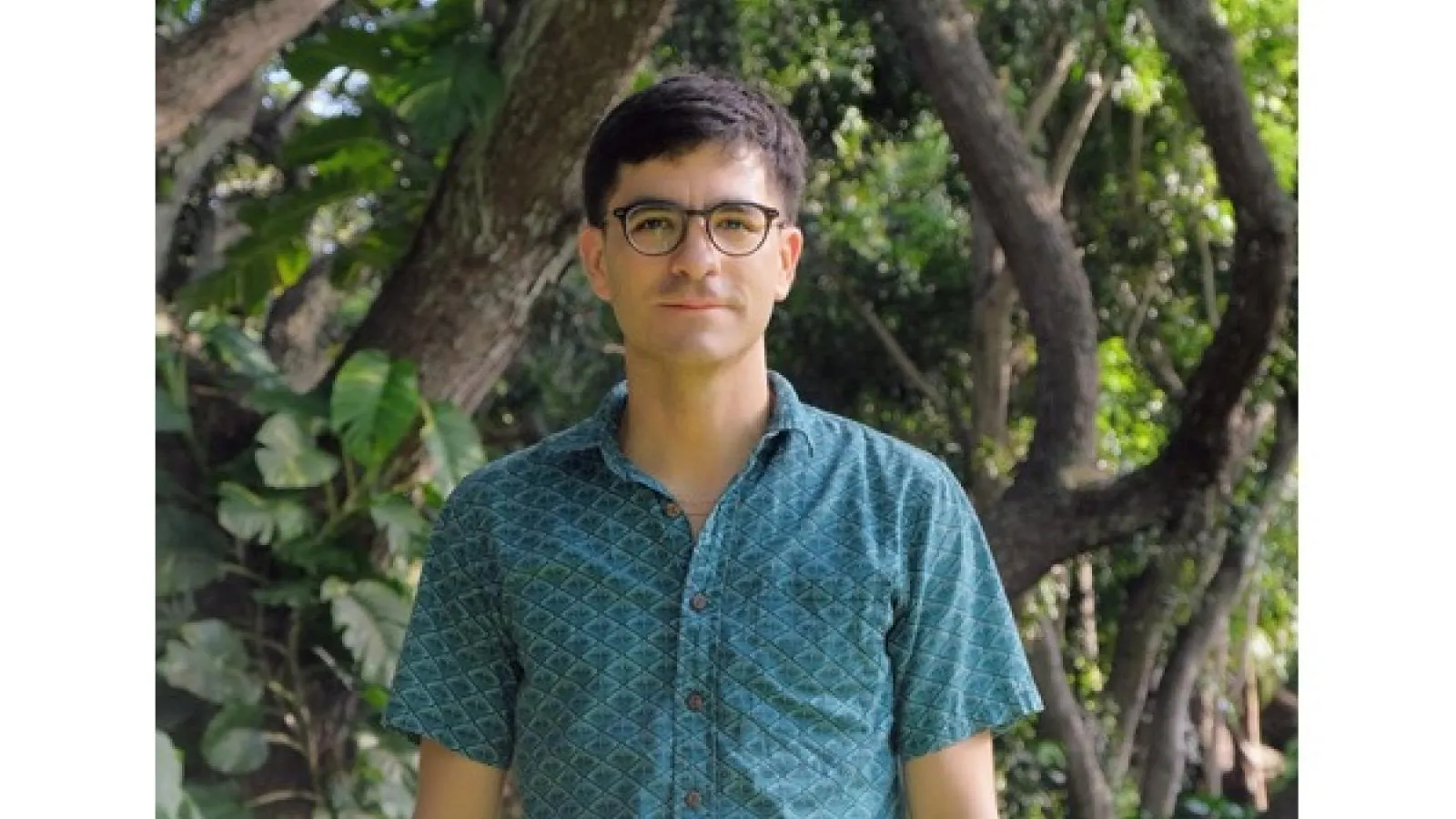 Picture of a man in a blue collored shirt with green trees in the background. 