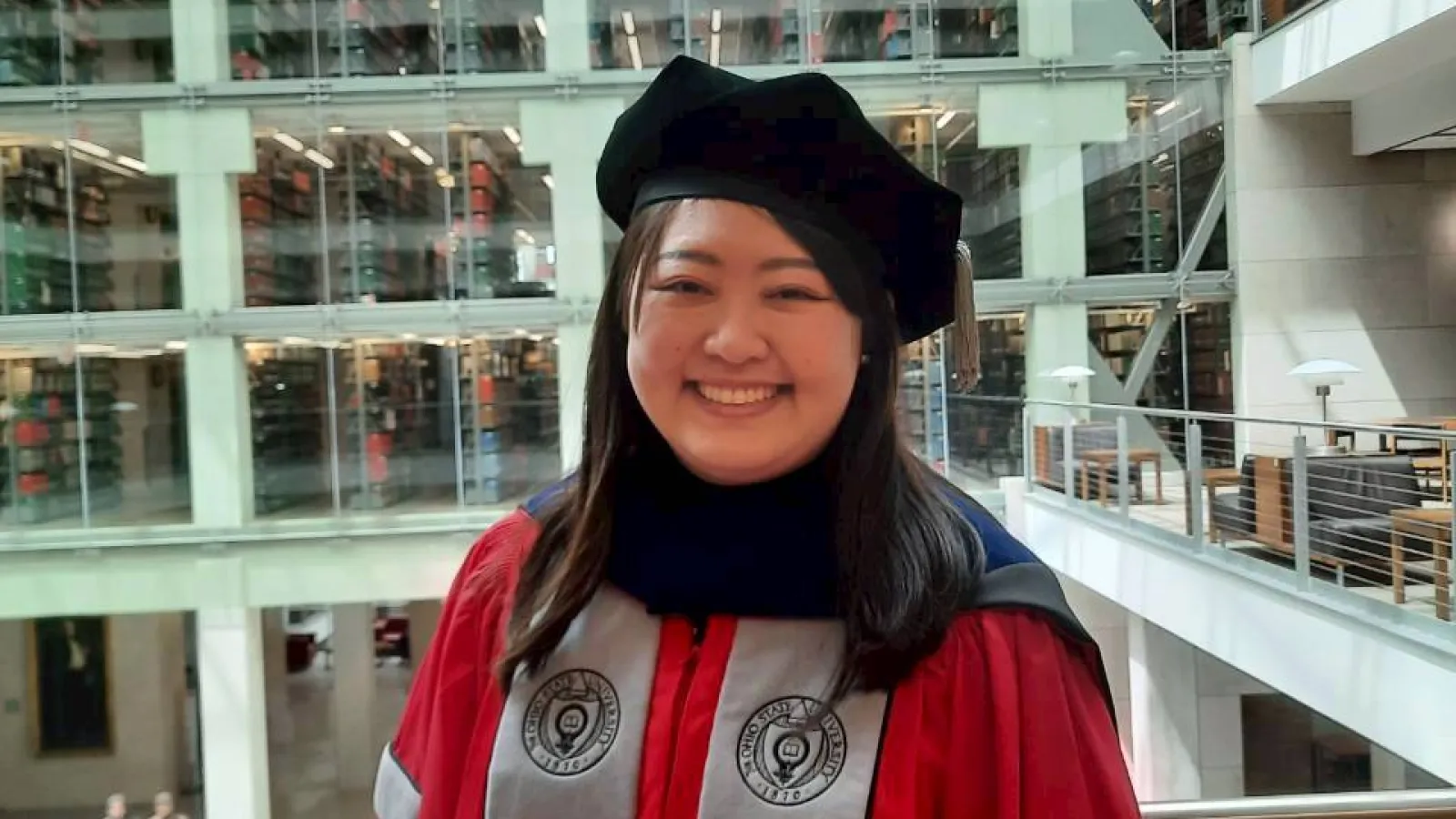 Graduate in red academic gown and black cap stands in a modern multi-story library with rows of books visible behind.