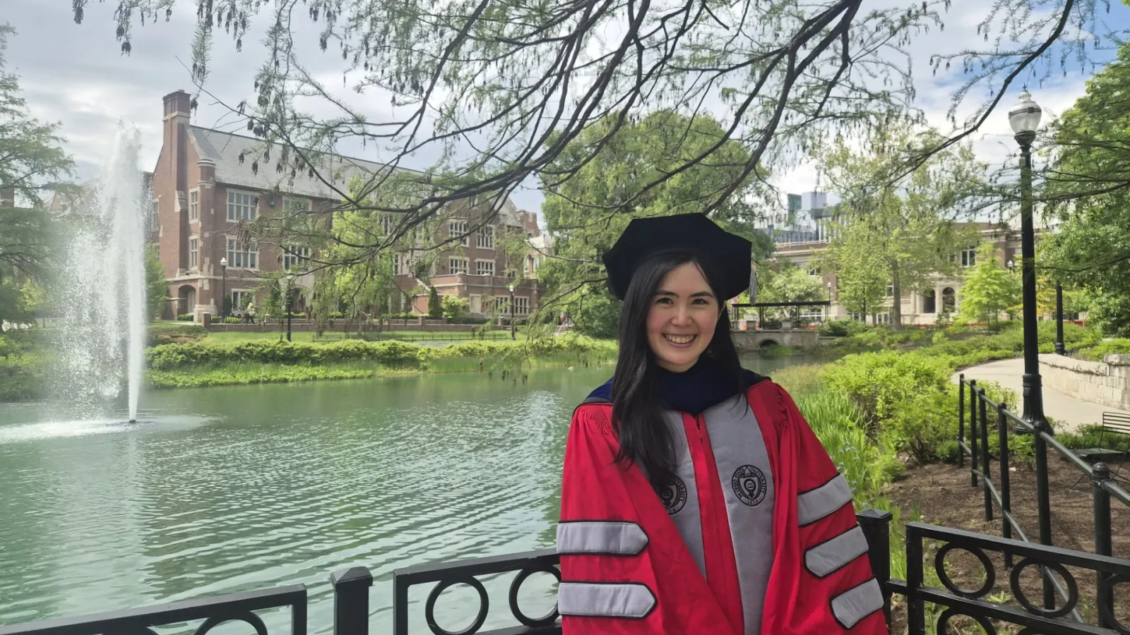 Person in a graduation gown by a lake with a fountain and historic buildings in the background.