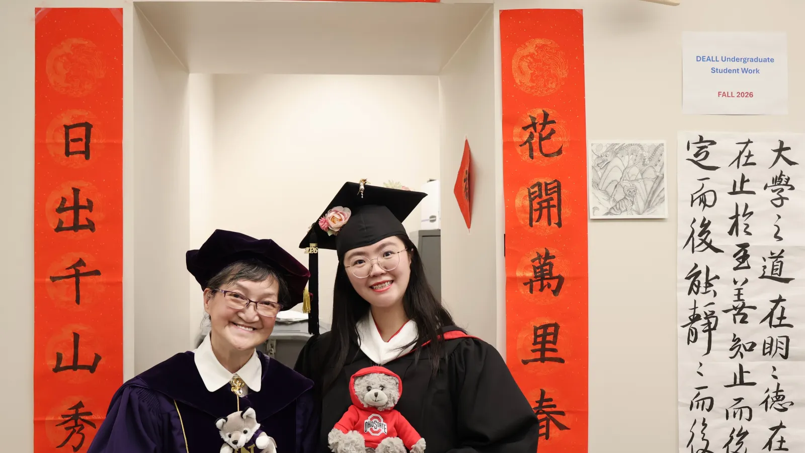 Two women in graduation gowns in front of a banner with Chinese writing. 