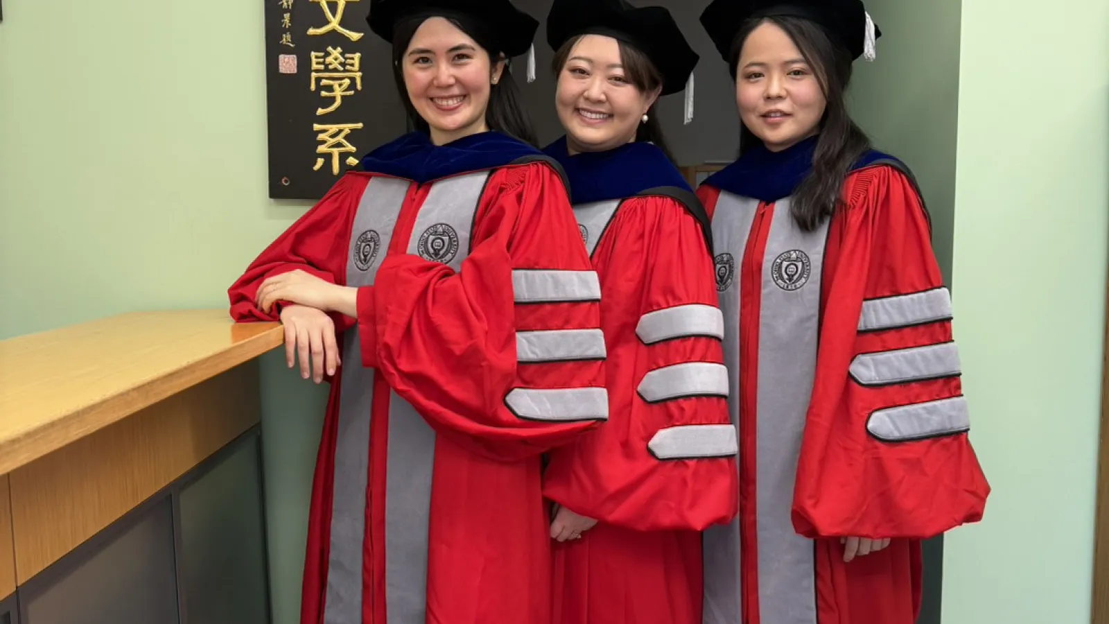  Three women in red and gray academic regalia with doctoral hoods and black caps stand indoors next to a sign with Chinese characters. 
