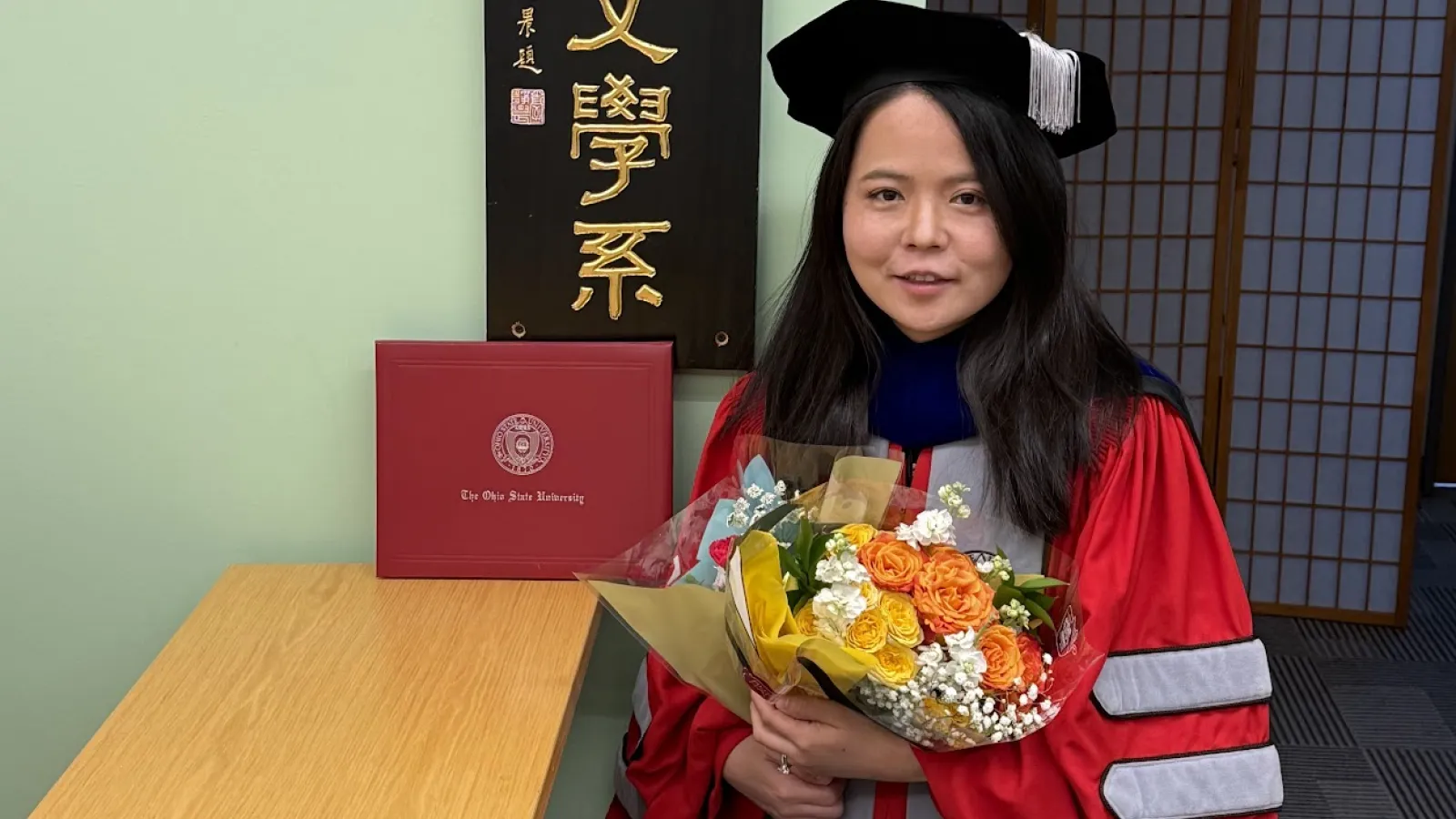  A woman in a red academic gown and cap holds a bouquet of flowers and stands next to a diploma and a sign with East Asian characters. 