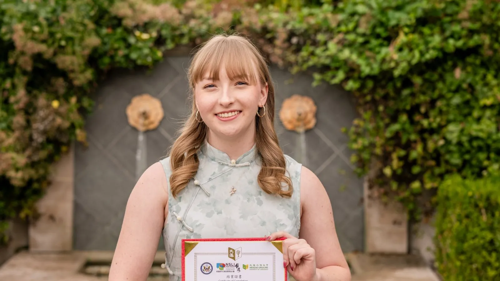 A woman proudly holds a certificate in front of a decorative fountain, smiling at the camera