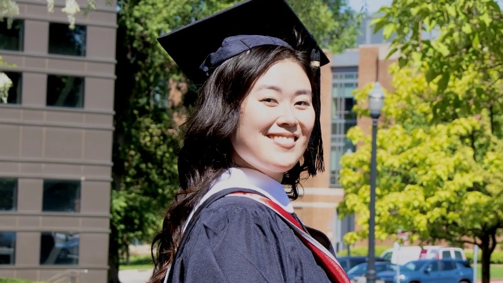 A young woman in graduation attire stands outdoors, smiling at the camera with trees and campus buildings in the background. 