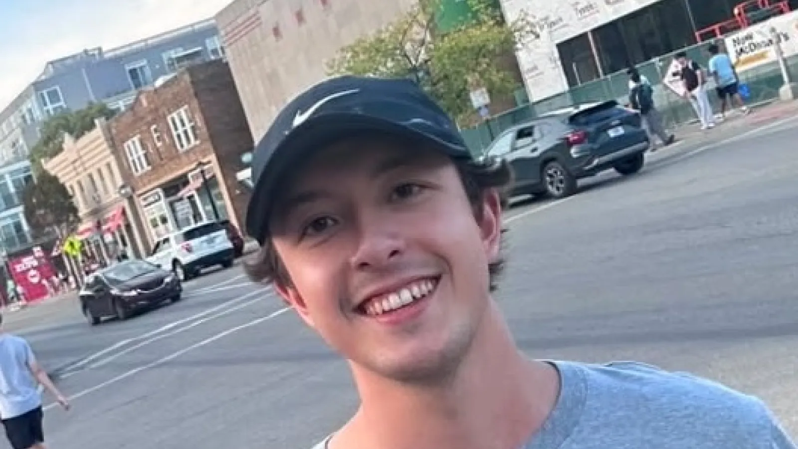 Man with dark baseball cap smiling, street scene in background 