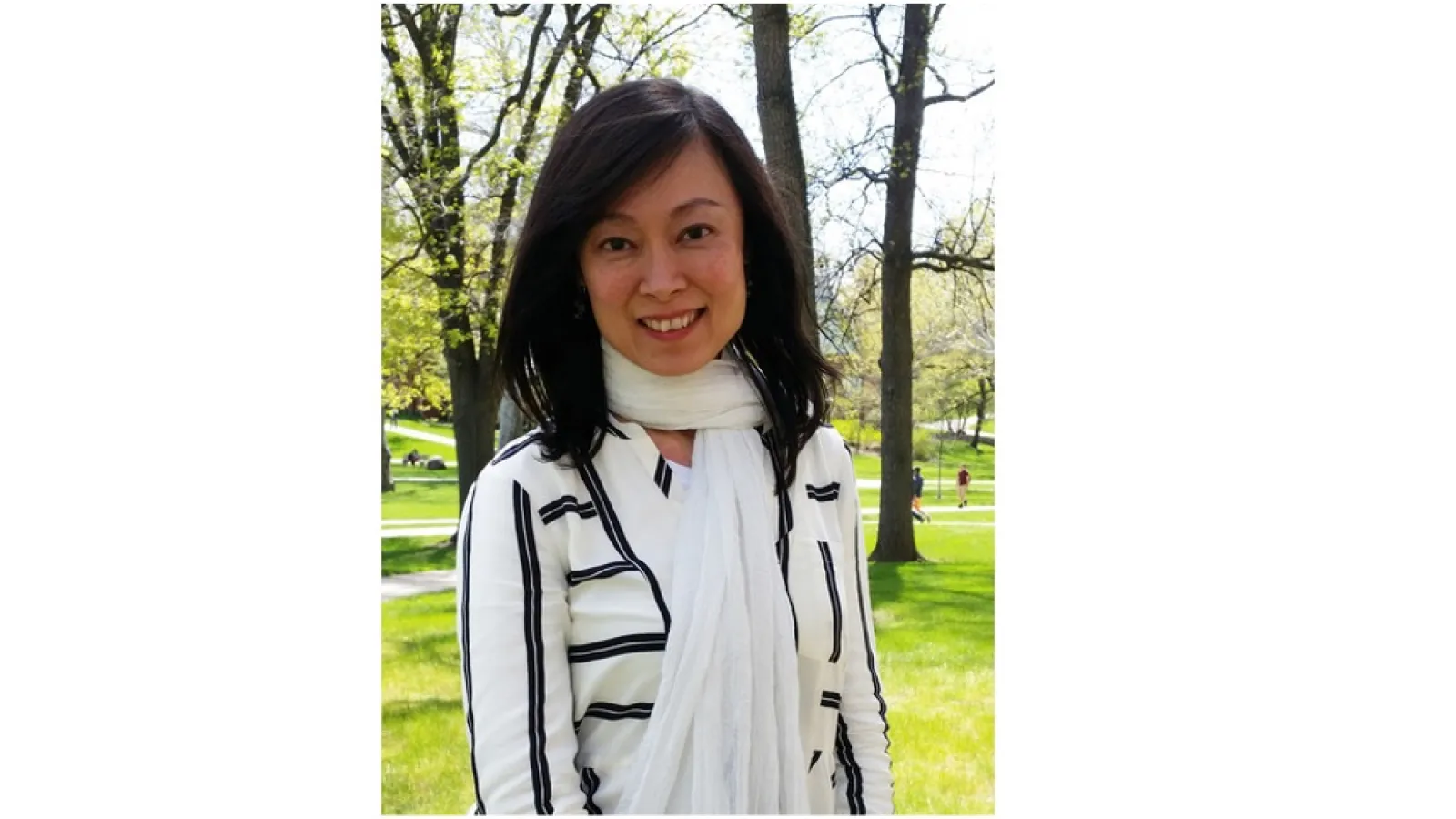 Woman with dark hair and a white scarf, smiling, and standing with trees behind her