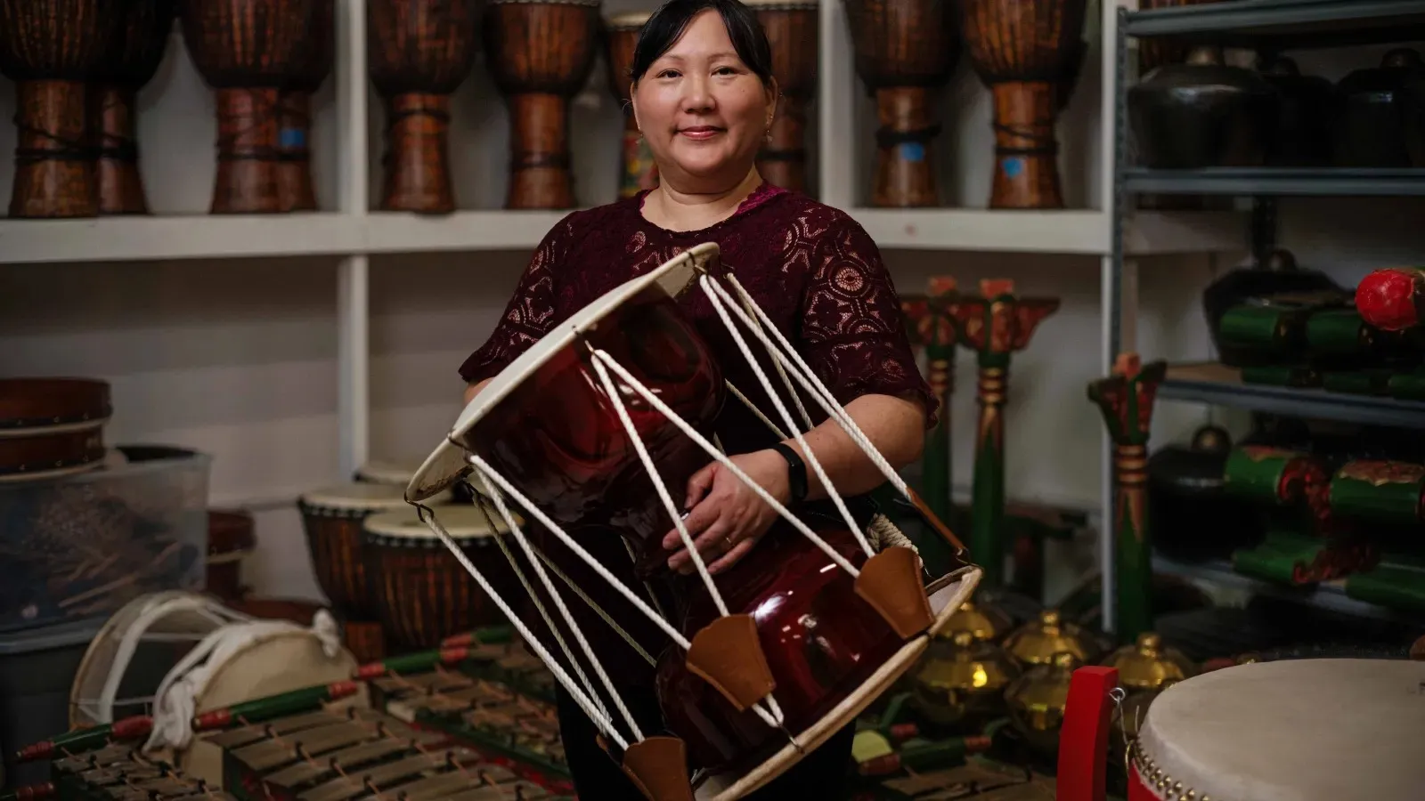 Person holding a large red rope-tension drum in a shop filled with percussion instruments.