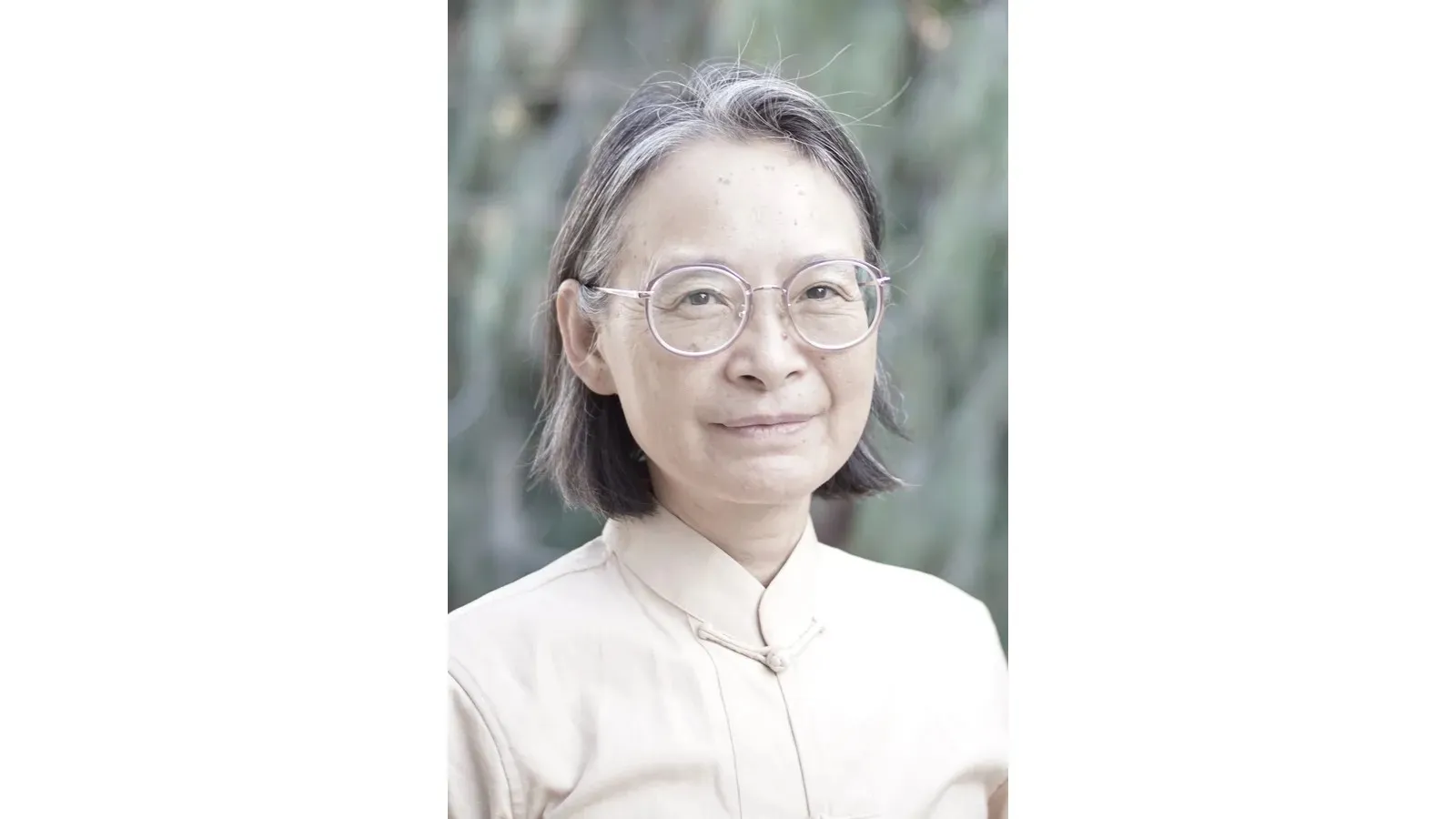 Close-up portrait of an older woman with short gray hair and round glasses, wearing a light-colored high-collared blouse.