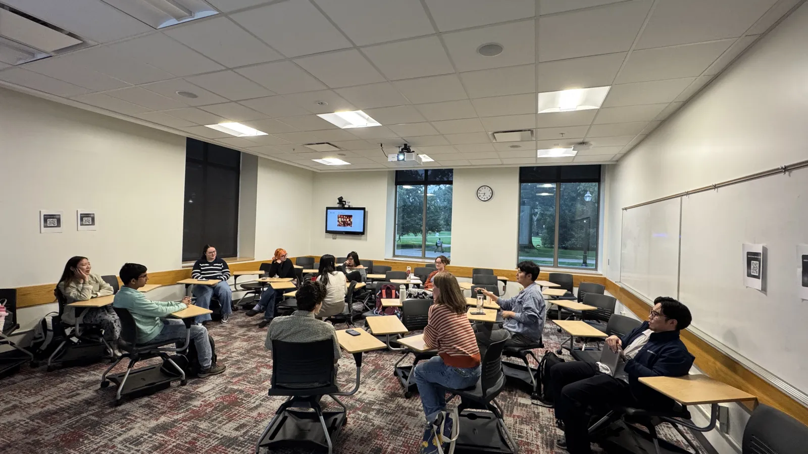 Students sit and converse in a classroom with desks, large windows, a ceiling-mounted projector, a wall display, and an analog clock.