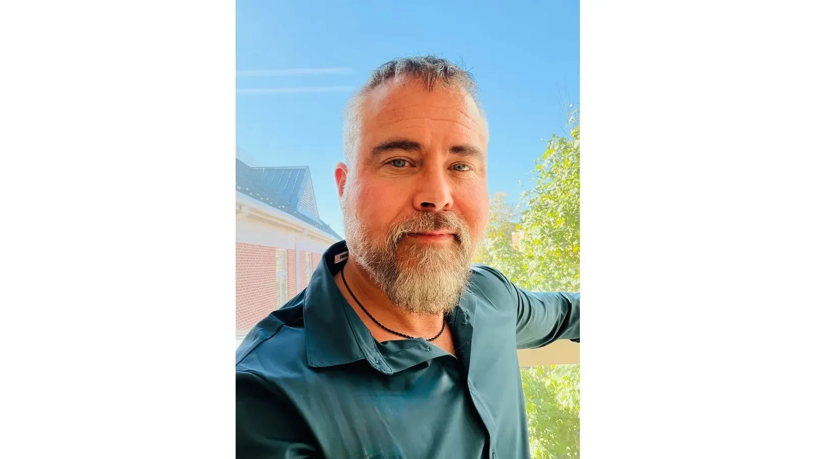 Close-up portrait of a bearded adult in a dark teal shirt standing outdoors beside a railing, with a brick building, blue sky, and leafy trees in the background.