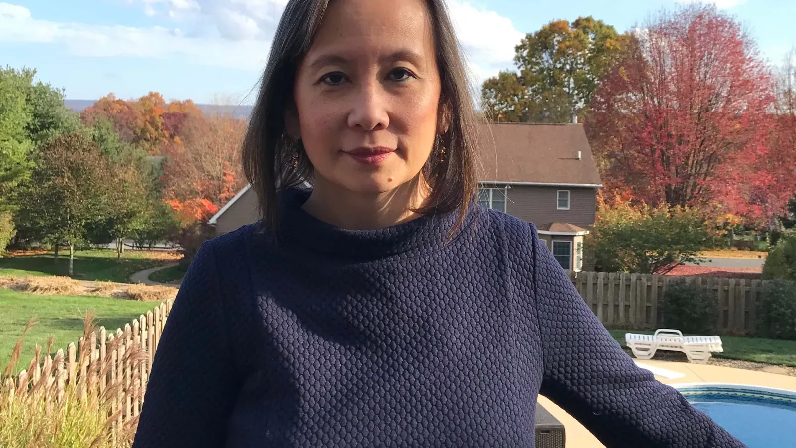 Woman in a navy textured top standing in a backyard surrounded by colorful autumn trees.