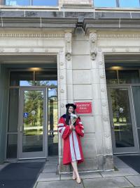 Woman in a graduation gown stands in front of Hagerty Hall, holding a diploma, with two glass doors and stone columns in the background.
