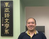 The image shows a person standing indoors next to a vertical black plaque with gold Chinese characters. The background features two walls, one painted in light green and the other in beige. The person is wearing a navy blue collared shirt with a yellow interior and is smiling at the camera. 