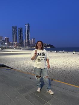  A young woman in a white t-shirt and jeans stands on a promenade by a sandy beach at dusk, with tall city buildings and lightsof Seoul in the background. 