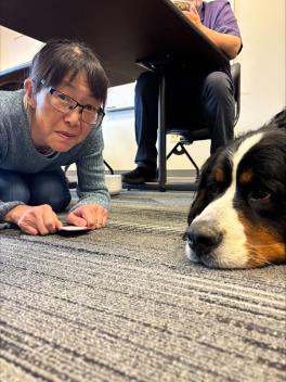 A woman crouches on a carpeted floor beside a large, relaxed dog. She holds a card and smiles slightly. The setting is a casual indoor space.