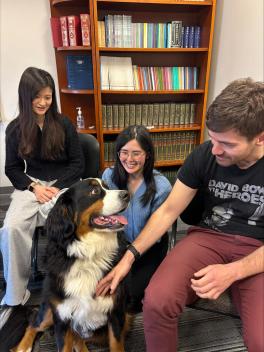 A large, fluffy dog sits happily among three smiling people in a room with a bookshelf. The scene is warm and friendly, conveying joy and companionship.
