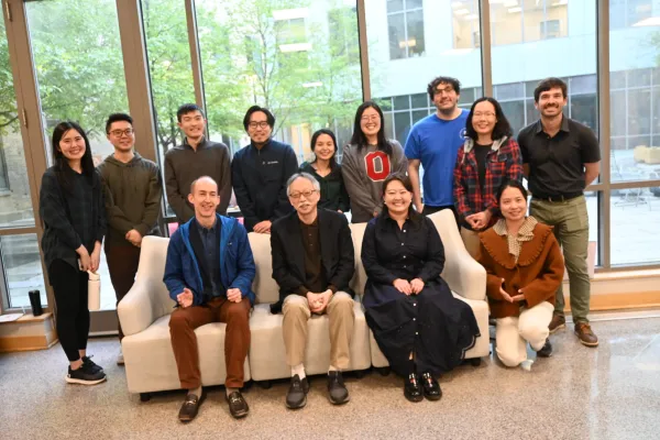  A group of thirteen people pose for a photo indoors, with three seated on a white couch and ten standing behind them in front of large windows. 