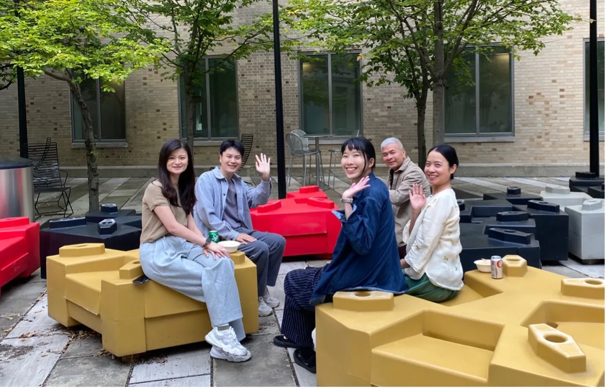 Five people seated on a bench in front of a building, engaged in conversation and enjoying the outdoors.  
