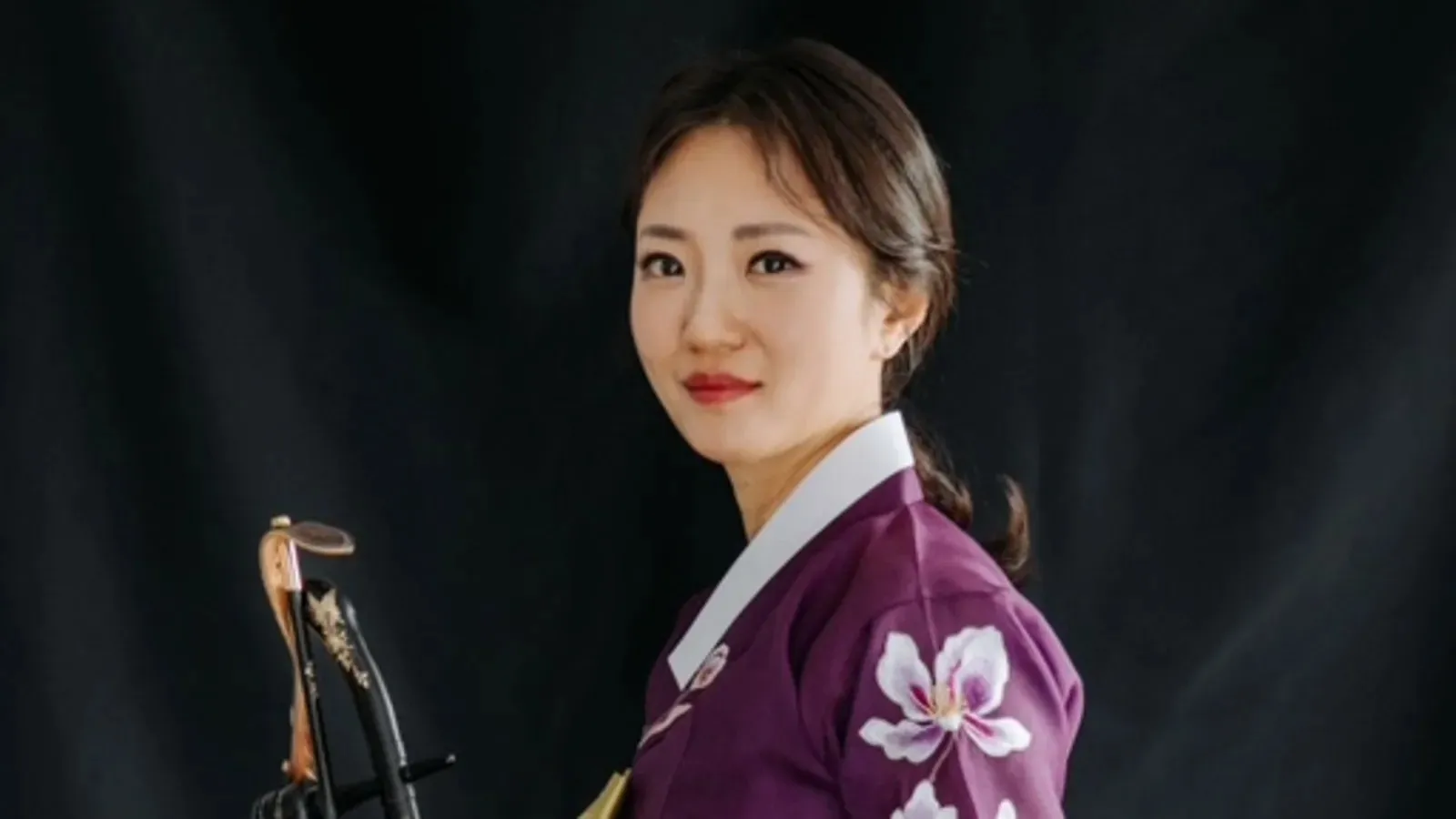 Smiling woman in a purple floral traditional garment holding a stringed instrument against a dark background.