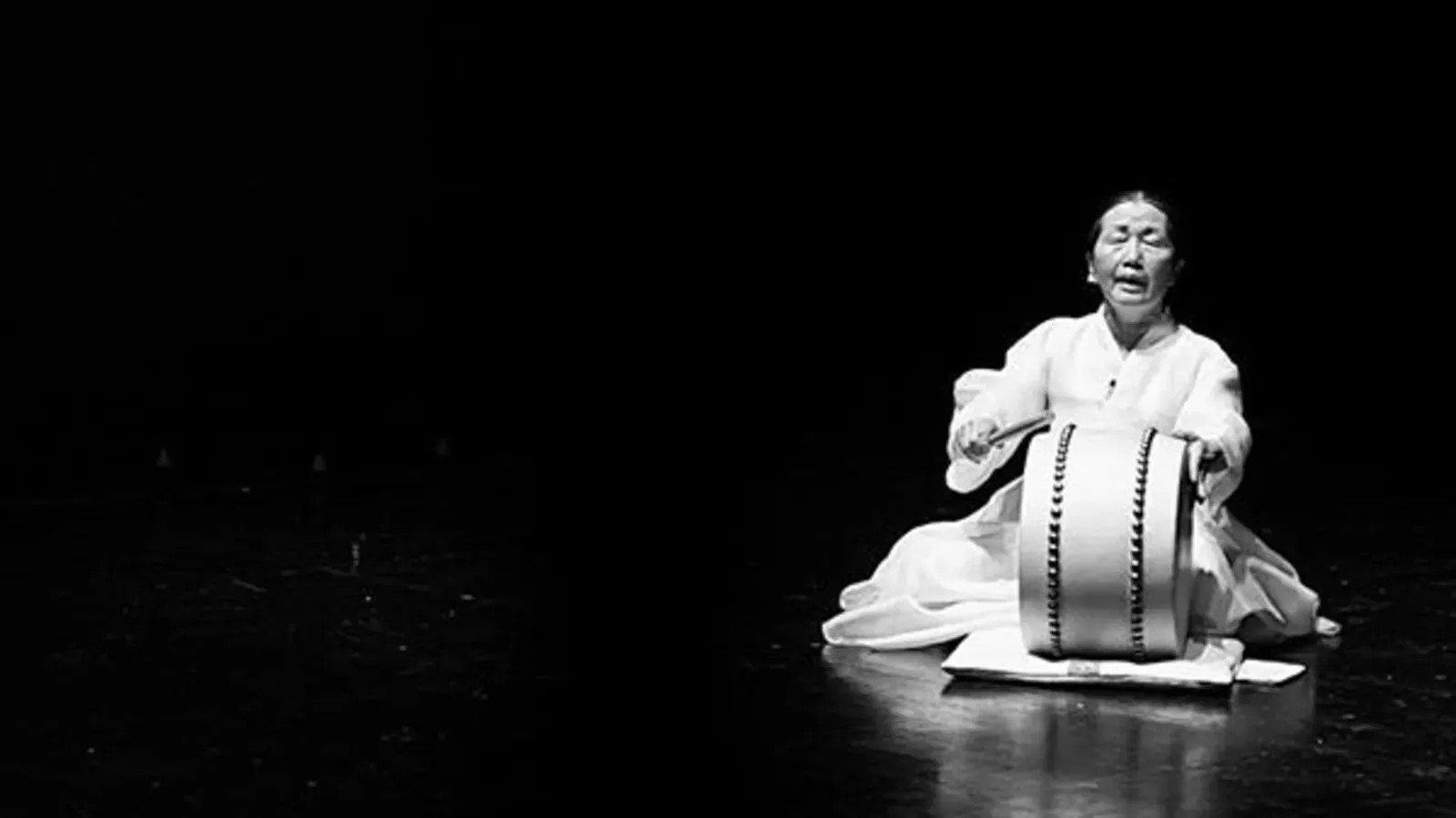 Black-and-white photograph of a seated performer in a light robe playing a large vertical drum on a dark stage.