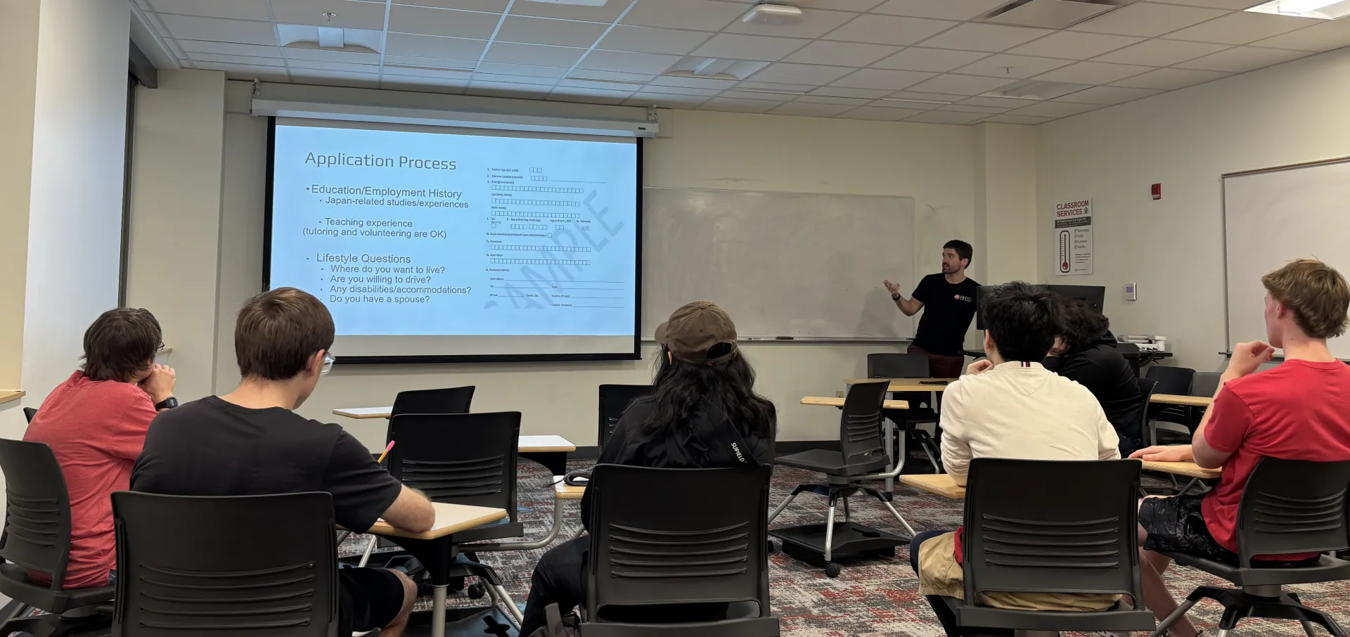 A man stands in front of a classroom next to a large, digital display, six students facing him in chairs