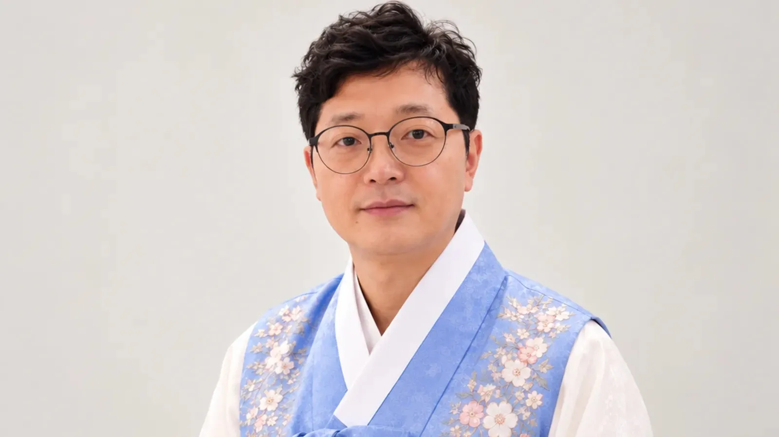 Portrait of a person wearing round glasses and a pale blue floral-embroidered vest over white clothing.