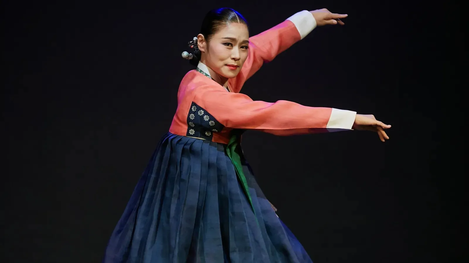 Dancer in a coral-pink top and full dark-blue skirt poses with both arms extended against a black background.