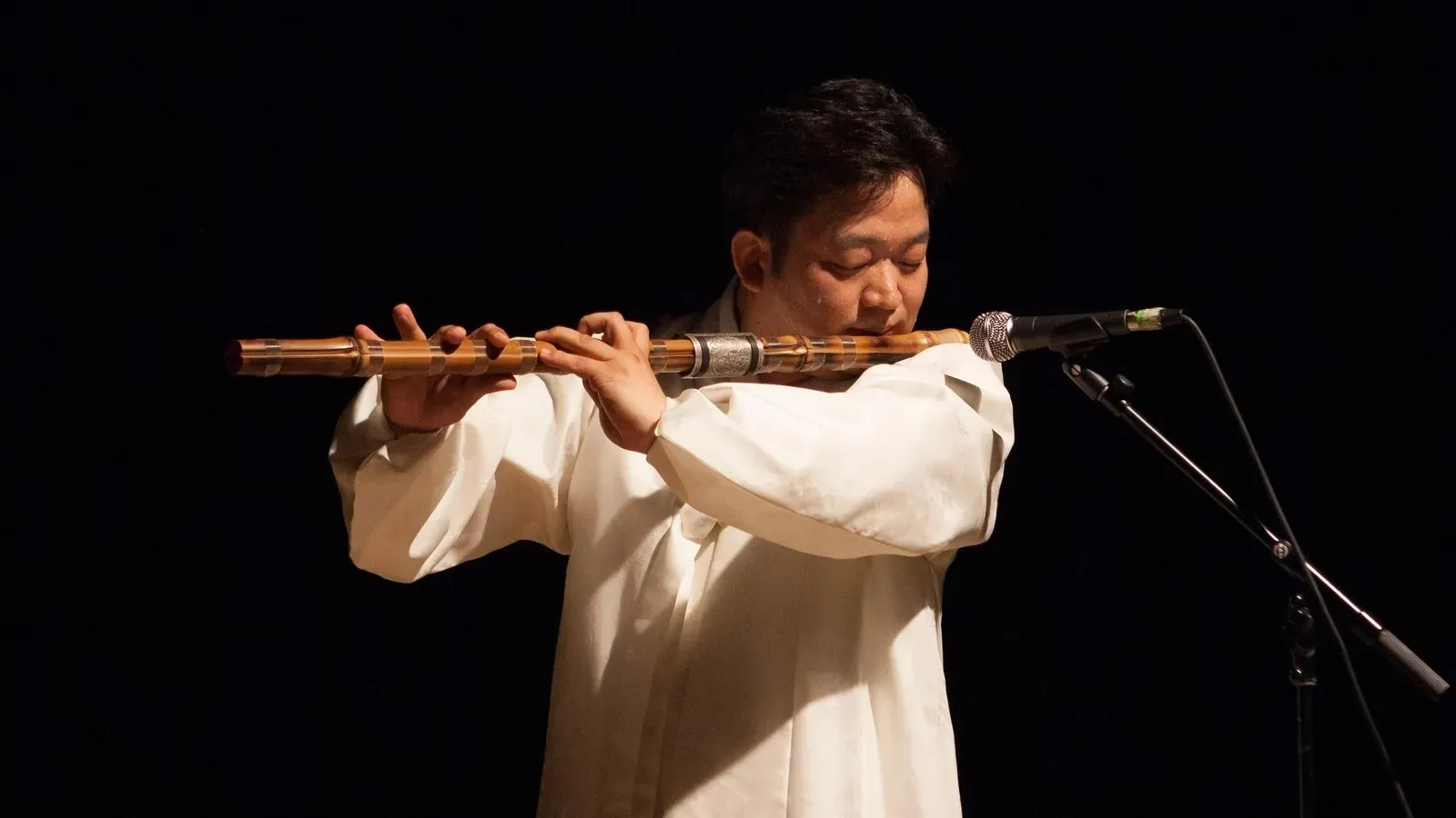 Musician playing a bamboo flute into a microphone on a dark stage.