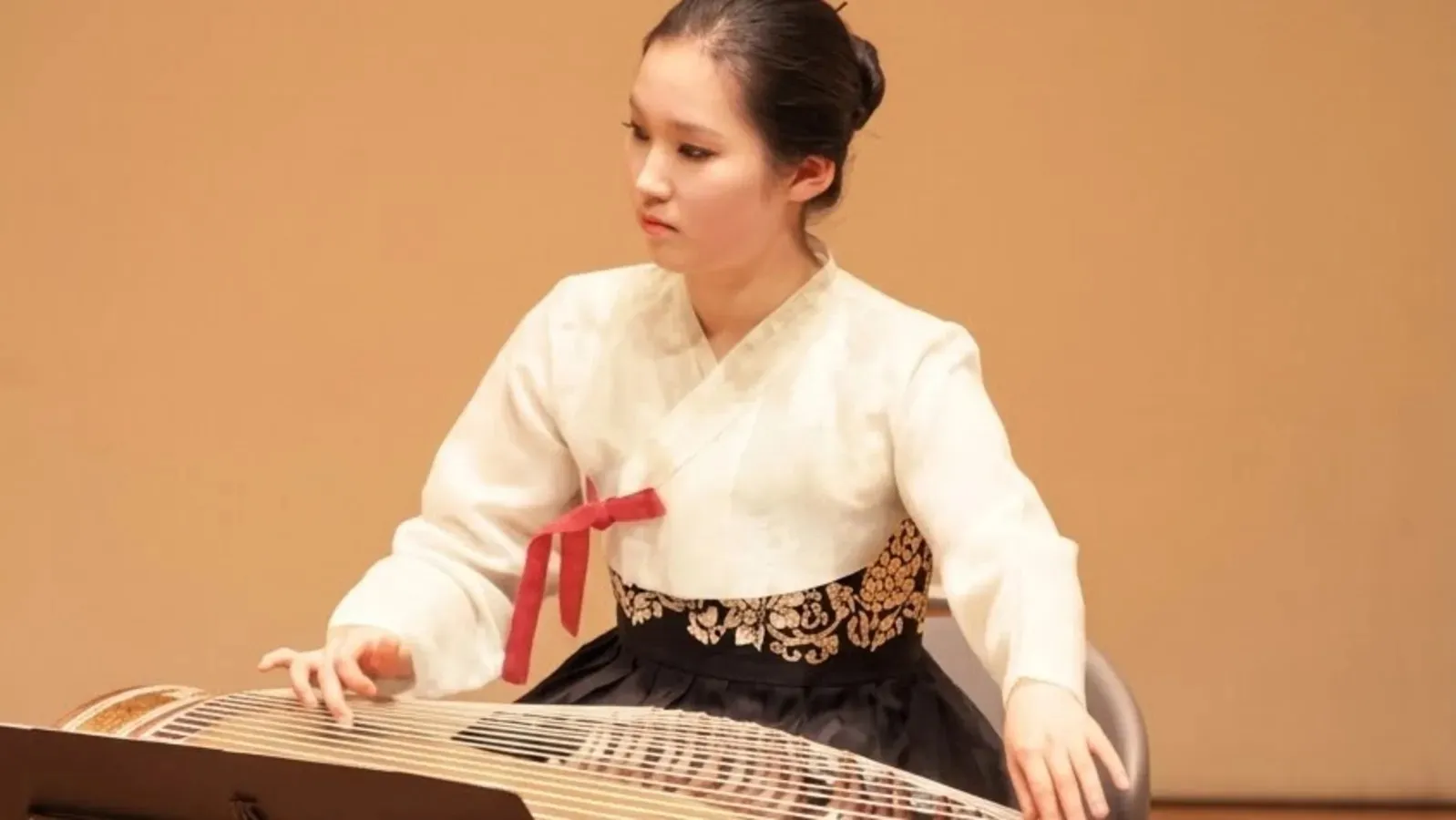 Woman in traditional clothing playing a large string instrument against a tan background.
