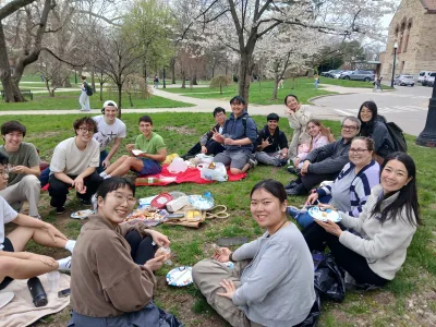  A group of people sit on blankets having a picnic in a park, smiling at the camera with food and trees visible around them. 